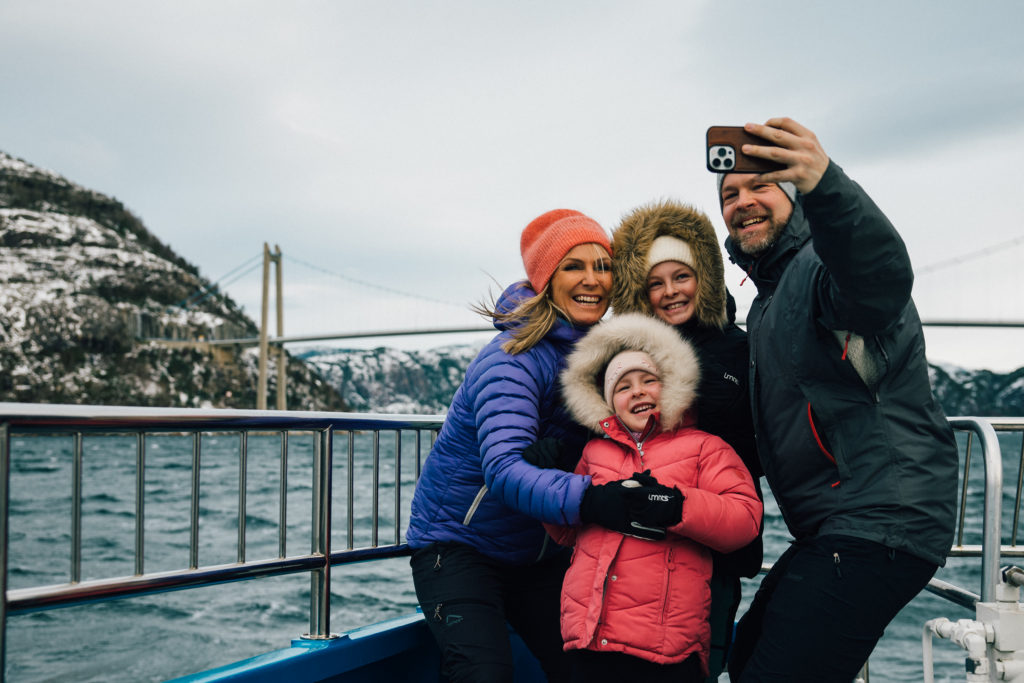 Familie tar selfie på et fjordcruise med fjell i bakgrunn og fjord samt en bro. Snø på fjellene.